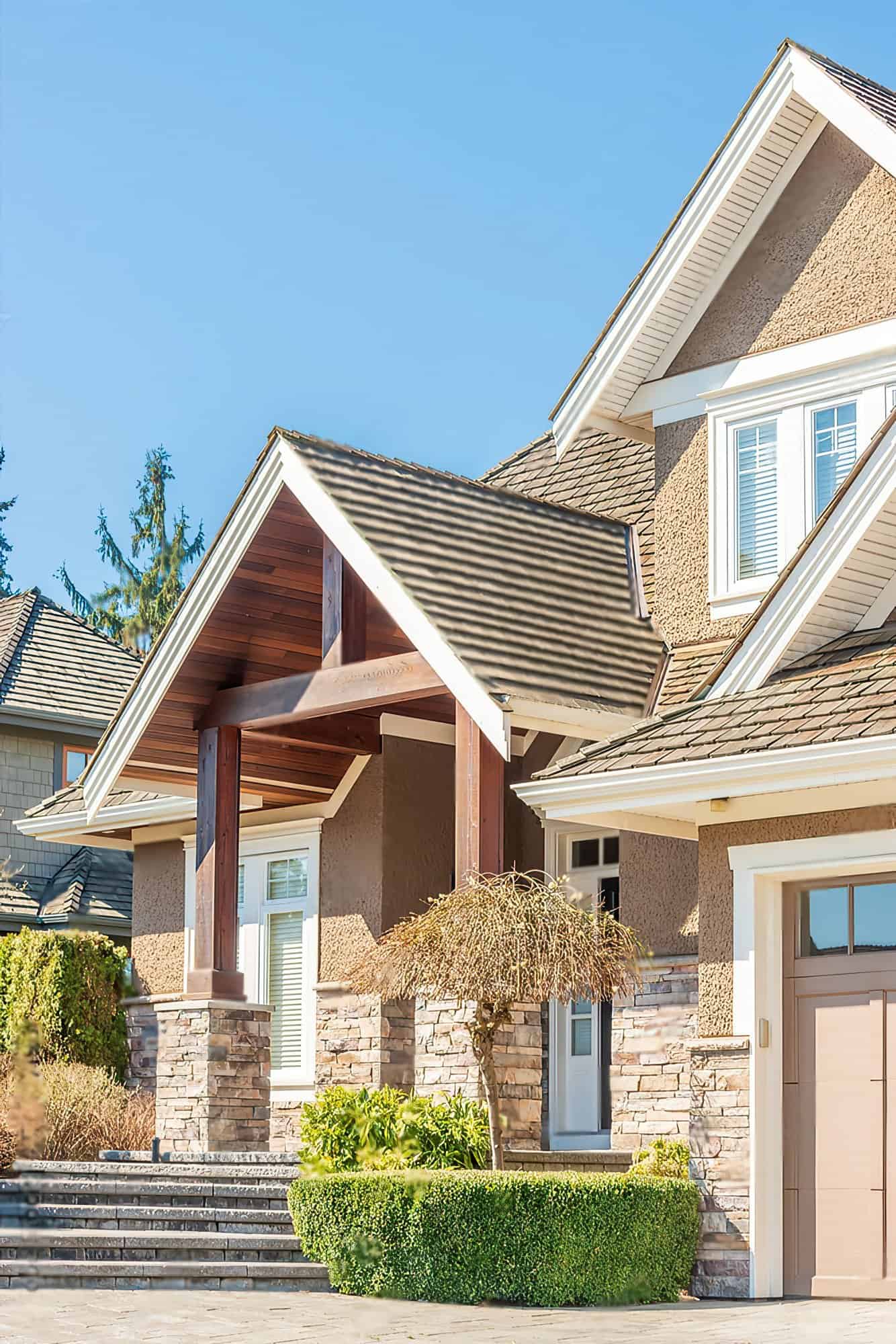 A large two-story house with a brown stucco exterior and a composite shingle roof. The house has a garage attached to the left side. The house also has a covered front porch with white columns.