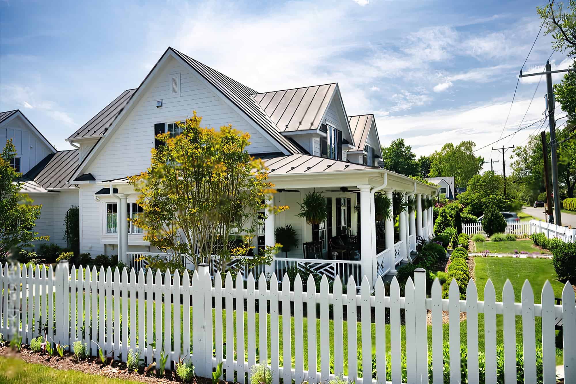 Single-story white house with a black picket fence in front of it. There are green bushes lining the front of the house, and a clear blue sky above it.