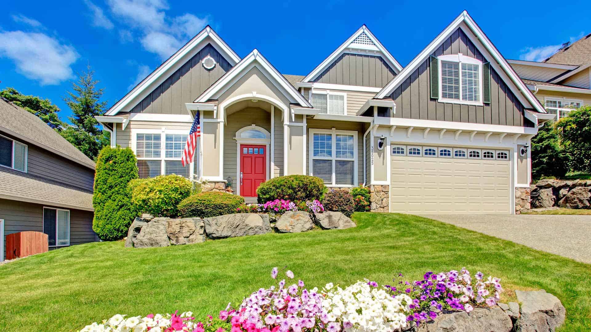 The house has a lush green lawn surrounding it, with flowers planted near the front door