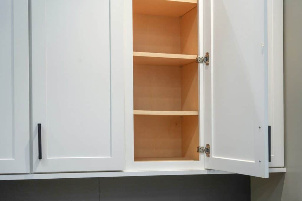 White-painted kitchen cabinets with open doors revealing natural wood interior shelves and metal hinges.