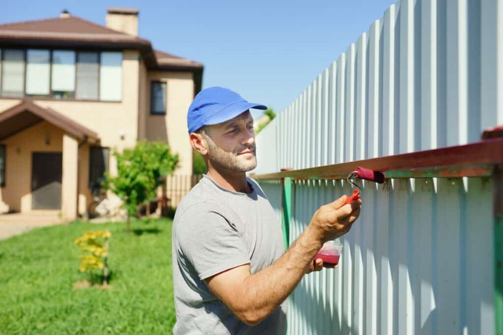 Painter applying stain to green corrugated metal fence panel with brush, residential home in background