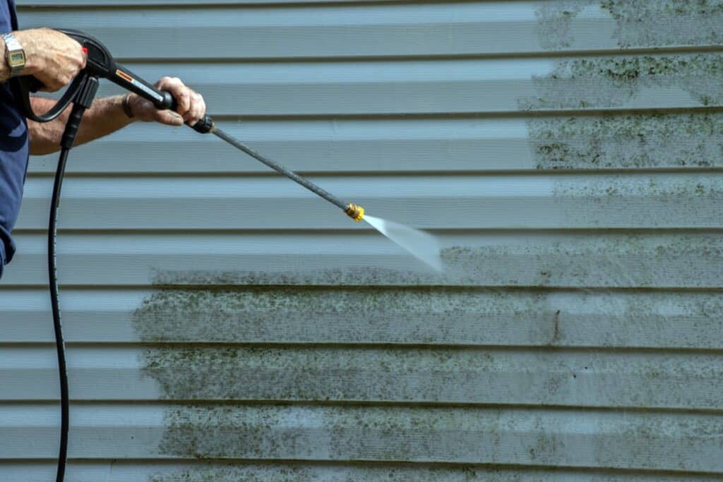 Worker pressure washing light blue horizontal vinyl siding with high-pressure spray, removing algae and dirt buildup